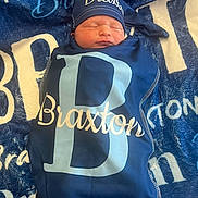 Braxton is registered to the contest to win money with this photo: newborn, baby, sleeping, blanket, hat, personalized, navy_blue, cozy, soft, wrapped, infant, child, resting, text, pattern, fabric, portrait, cute, peaceful, indoors