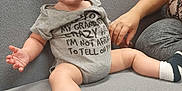 Levi joined the competition — help win amazing prizes! baby, child, onesie, socks, couch, indoor, person, hand, seated, curious, expression, casual_clothing, support, adult, furniture, grey, text_on_clothing, legs, face, blue_eyes