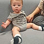baby, child, onesie, socks, couch, indoor, person, hand, seated, curious, expression, casual_clothing, support, adult, furniture, grey, text_on_clothing, legs, face, blue_eyes