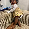 baby, child, smile, happy, curly_hair, leopard_print, dress, diaper, couch, tattoo, arm, hand, indoor, furniture, foot, skin, person, infant, climbing, playful
