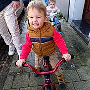 Owen participe au concours pour gagner de l'argent avec cette photo : child, toddler, tricycle, sidewalk, outdoor, smiling, person, jacket, shoes, pavement, urban, building, window, door, hand, toy, vehicle, casual_clothing, daytime, play