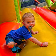 Maël a rejoint le concours — aidez-le/la à gagner de superbes lots ! blue_shirt, blue_socks, building, child, climbing, fun, grass, happy, inflatable_bounce_house, kids, orange, outdoor, pacifier, play, playground, red, summer, toddler, trees, yellow