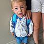 Maël a rejoint le concours — aidez-le/la à gagner de superbes lots ! backpack, blue_shirt, casual_clothing, child, closeup, curious_expression, floor, flooring, hand, indoor, legs, light_hair, person, shoes, shorts, sibling, sneakers, standing, toddler, white_jacket