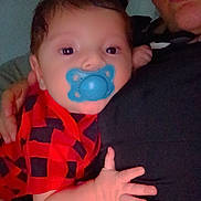 Milo is registered to the contest to win money with this photo: baby, pacifier, red_clothing, checkered_pattern, adult, face, hand, close_up, indoor, portrait, infant, child, resting, casual, person, black_clothing, skin, holding, comfort, family