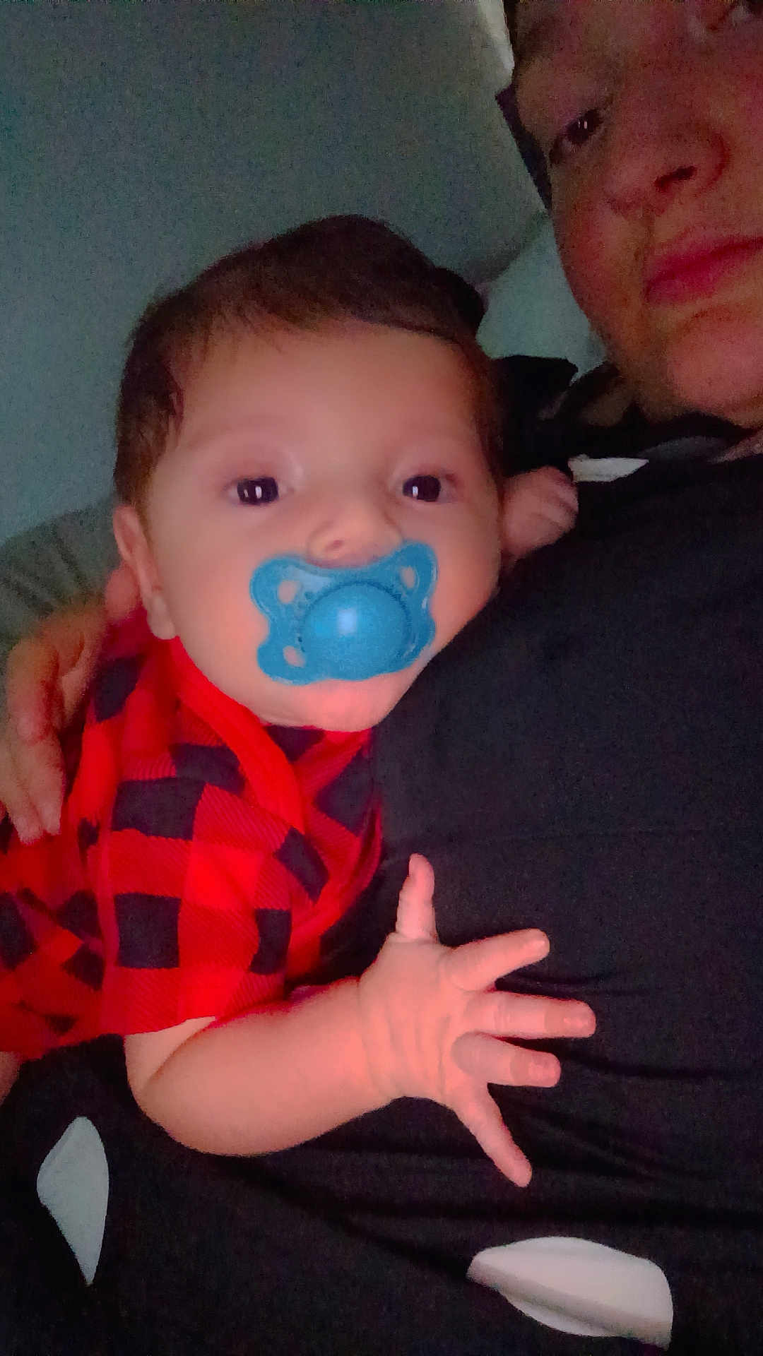 Milo is registered to the contest to win money with this photo: baby, pacifier, red_clothing, checkered_pattern, adult, face, hand, close_up, indoor, portrait, infant, child, resting, casual, person, black_clothing, skin, holding, comfort, family