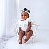 baby, child, smiling, white_outfit, headband, flower_crown, chair, sitting, cute, portrait, indoor, curtains, white_background, infant, happy, toddler, fashion, adorable, studio, young_child