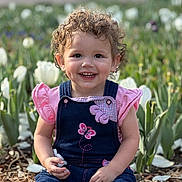 Nova is registered to the contest to win money with this photo: child, toddler, smiling, curly_hair, pink_clothing, denim_overalls, butterfly_patch, flower_garden, white_tulips, outdoor, spring, nature, plants, greenery, sunlight, happy, portrait, sitting, cute, young_child