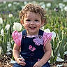 child, toddler, smiling, curly_hair, pink_clothing, denim_overalls, butterfly_patch, flower_garden, white_tulips, outdoor, spring, nature, plants, greenery, sunlight, happy, portrait, sitting, cute, young_child