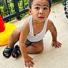toddler, child, sitting, white_tank_top, gray_shorts, black_slides, nike, outdoor, concrete_floor, plants, fence, curious_expression, barefoot, summer_clothing, young_child, playful, casual, daylight, person, portrait