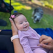 Amaya joined the competition — help win amazing prizes! baby, smiling, infant, pink_bow, purple_clothing, hand, outdoor, grass, dog, blurred_background, person, watch, nature, happy, portrait, toddler, cute, holding, skin, playful