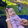 baby, smiling, infant, pink_bow, purple_clothing, hand, outdoor, grass, dog, blurred_background, person, watch, nature, happy, portrait, toddler, cute, holding, skin, playful