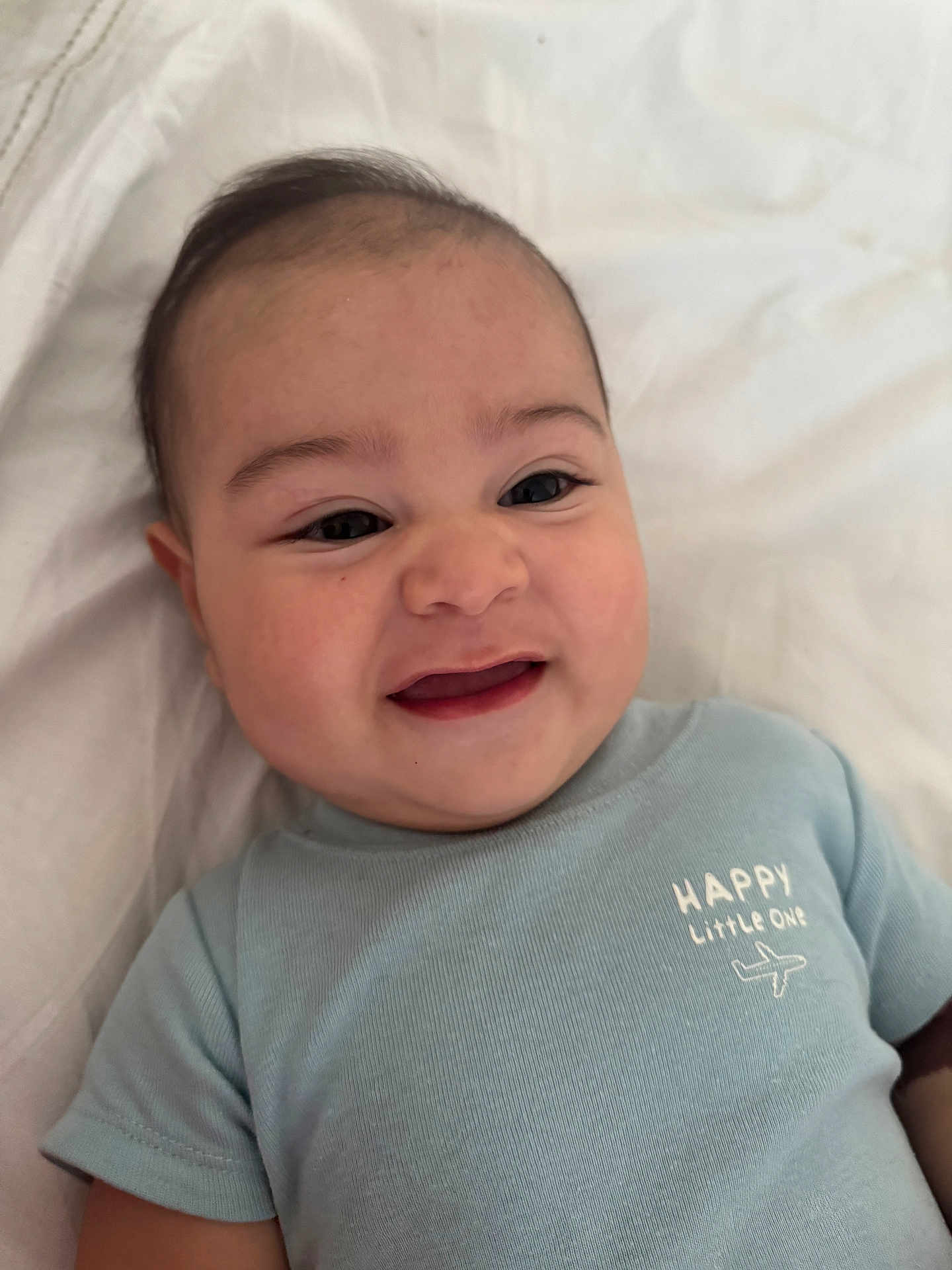 Maximiliano is registered to the contest to win money with this photo: baby, smiling, infant, child, cute, portrait, blue_shirt, text_on_clothing, happy, lying_down, face, eyes, mouth, skin, head, short_hair, blanket, soft_texture, indoors, close_up