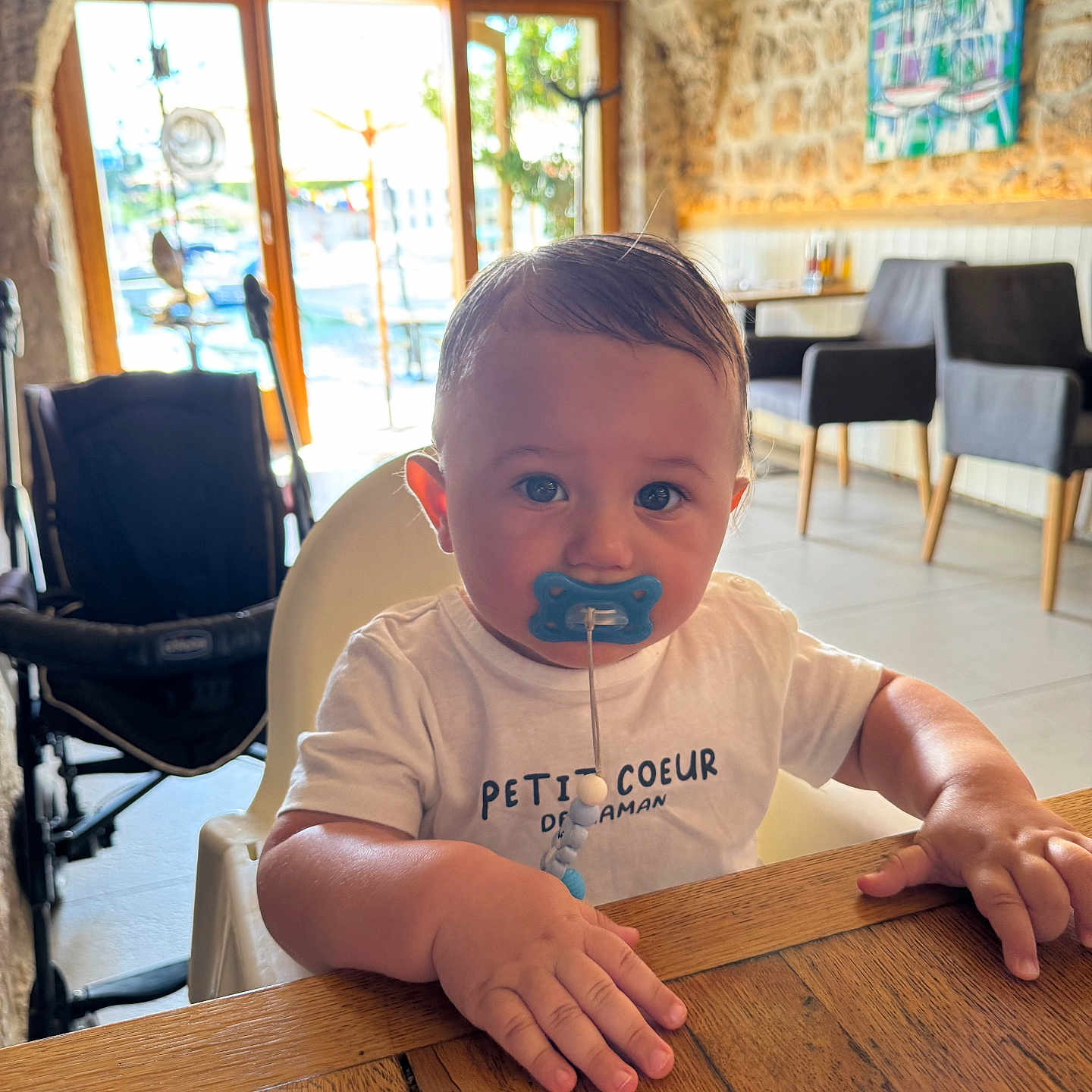 Nahyl a rejoint le concours — aidez-le/la à gagner de superbes lots ! baby, blue_pacifier, cafe, chair, child, cute, daylight, face, furniture, hands, highchair, indoor, pacifier, portrait, stone_wall, stroller, toddler, white_shirt, window, wooden_table