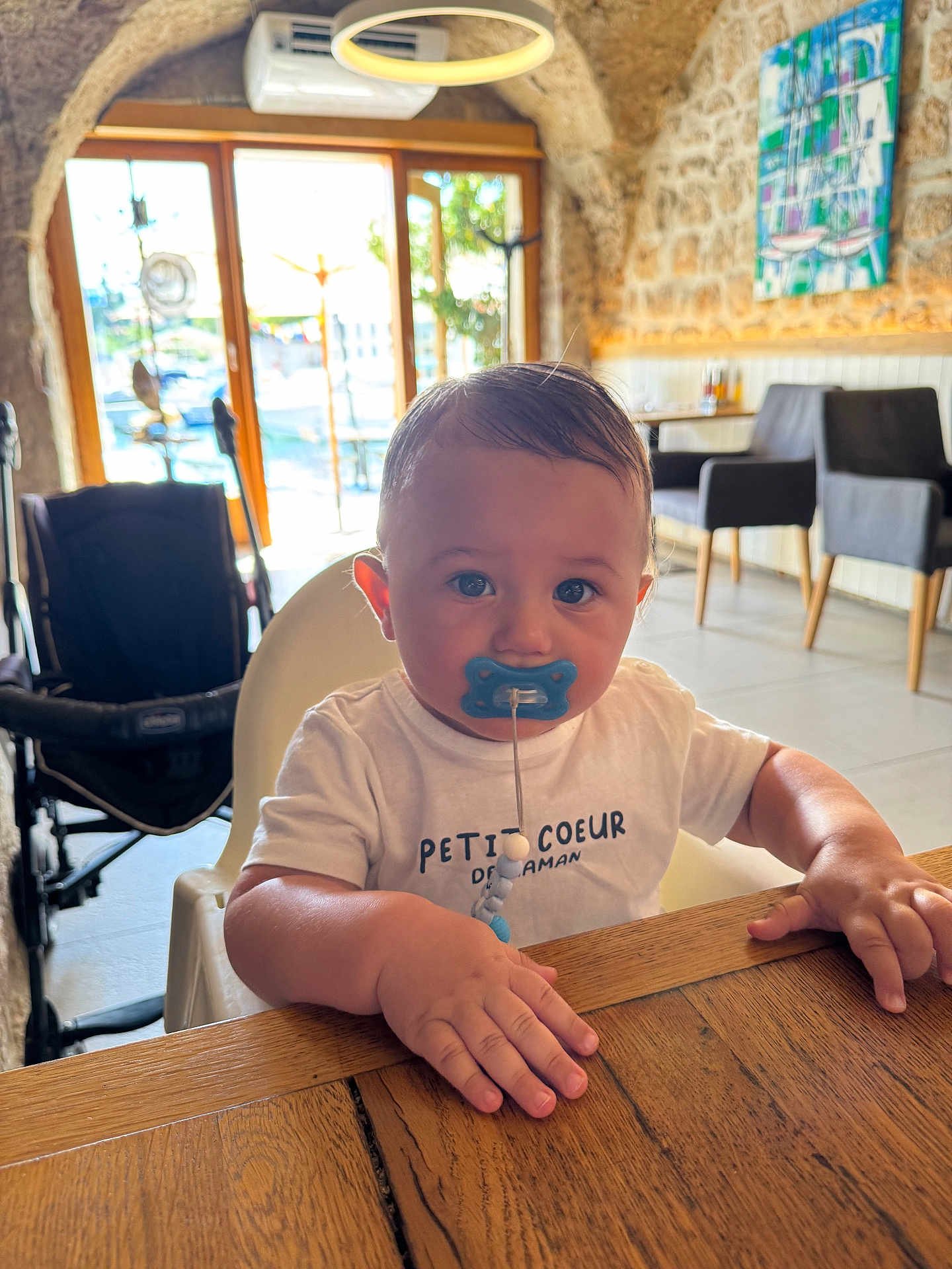 Nahyl a rejoint le concours — aidez-le/la à gagner de superbes lots ! baby, child, pacifier, highchair, wooden_table, indoor, cafe, stone_wall, window, daylight, white_shirt, blue_pacifier, face, hands, chair, stroller, furniture, portrait, cute, toddler