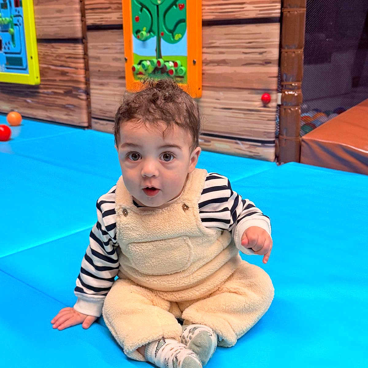 Nahyan participe au concours pour gagner de l'argent avec cette photo : baby, ball, child, colorful_panel, curly_hair, expression_surprised, eyes, face, fuzzy_overalls, indoor_play_area, play_mat, portrait, sitting, socks, soft_texture, striped_shirt, toddler, toys, wood_panel_wall, young_child