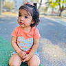 toddler, child, outdoor, playground, kneeling, curly_hair, orange_shirt, heart_design, shorts, daylight, trees, background_blur, expression, casual_clothing, young_child, skin, face, person, nature, cute