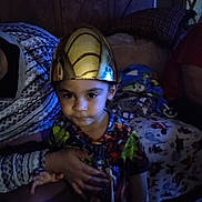 Jayden is registered to the contest to win money with this photo: child, helmet, golden_helmet, colorful_shirt, person, indoor, blanket, pillow, hand, adult, seated, furniture, curious, dim_lighting, home, portrait, young, face, couch, cozy