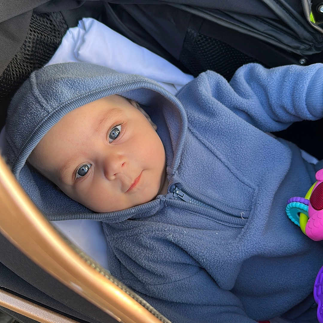 Noah Cristian is registered to the contest to win money with this photo: baby, baby_toy, blue_hoodie, bundle, child, closeup, curious, cute, expression, eyes, face, fleece, hood, infant, outdoor, portrait, seated, stroller, toy, warm_clothing