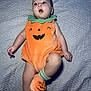 baby, infant, child, pumpkin_costume, orange_clothing, plush, sock, footwear, blanket, bed, lying_down, cute, expression, face, head, skin, person, indoor, soft_texture, holiday_costume