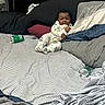 baby, child, pajamas, bed, pillow, striped_bed_sheet, soda_can, smile, indoor, sleepwear, messy_bed, person, headboard, fabric, cute, infant, relaxing, black_hair, lying_down, cozy