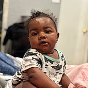 Dream is registered to the contest to win money with this photo: baby, child, curly_hair, shirt, shark_pattern, blanket, indoor, portrait, sitting, cute, infant, diaper, expression, skin, hand, face, eyes, person, young, cozy