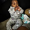 Dream is registered to the contest to win money with this photo: baby, child, pajamas, grey_clothing, sitting, bed, striped_bedspread, baby_wipes, plush_toy, fingers, curious, indoors, dark_background, cute, face, hands, foot, person, infant, cozy