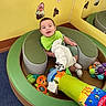 Jayden joined the competition — help win amazing prizes! baby, child, toys, green_bib, circular_seat, mirror, yellow_wall, carpet, stuffed_toy, plush_toy, colorful, smiling, indoor, play_area, infant, happy, seated, soft_furniture, reflection, cute