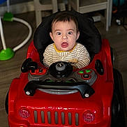 Jayden is registered to the contest to win money with this photo: baby, child, toy_car, indoor, surprised_expression, striped_clothing, flooring, chair, table, plastic_toys, furniture, playroom, head, face, person, seat, footwear, expression, young_child, playtime