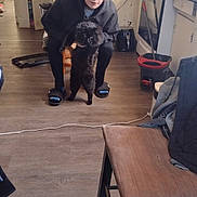 Savage is registered to the contest to win money with this photo: person, cat, indoor, kitchen, floor, chair, slippers, hoodie, black_cat, wooden_floor, household_items, appliance, mop, bucket, young_person, furniture, casual, pet, curly_hair, looking_at_camera