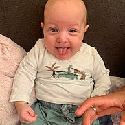 Raphaël a rejoint le concours — aidez-le/la à gagner de superbes lots ! baby, tongue_out, smiling, dinosaur_shirt, green_pants, couch, blanket, hand, playful, infant, cute, indoors, person, clothing, sitting, expression, human, skin, face, happy