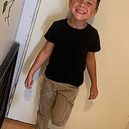 Lucas participe au concours pour gagner de l'argent avec cette photo : child, boy, smiling, black_tshirt, cargo_pants, indoor, door, floor_tiles, plant, happy, pose, footwear, sneakers, casual_clothing, young, playful, person, wall, standing, light
