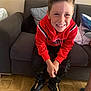 child, boy, smile, red_hoodie, black_pants, couch, living_room, pillow, floor, tile_floor, sitting, happy, casual_clothing, indoor, person, footwear, nike_shoes, hair, face, portrait
