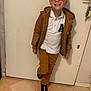 child, boy, glasses, brown_jacket, white_polo_shirt, mustard_pants, black_shoes, indoor, tiled_floor, door, smiling, standing, casual_clothing, pose, short_hair, wall, keys, happy, person, portrait