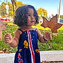 child, toddler, curly_hair, dress, leaves, autumn, outdoor, park, sidewalk, grass, tree, blue_sky, daylight, playful, nature, holding, person, cute, colorful, happy