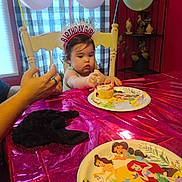 Harper is registered to the contest to win money with this photo: adult_hand, baby, balloons, birthday, birthday_party, cake, celebration, child, cupcake, disney_princesses, eating, frosting, high_chair, indoors, party_hat, plate, portrait, smiling, table, tablecloth