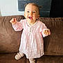brown_couch, child, couch, cute, daisies, face, hair, happy, home, indoor, joyful, person, pigtails, pink_dress, shoes, smiling, standing, texture, toddler, wall