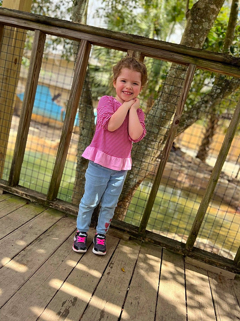 Briar is registered to the contest to win money with this photo: botany, fence, fun, grass, happy, jacket, joy, leg, leisure, mammal, morning, pattern, people_in_nature, person, pink, plant, recreation, smile, toddler, tree