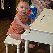 Hazelmarie is registered to the contest to win money with this photo: child, colorful_background, cute, floor, furniture, home, indoor, musical_instrument, orange_clothing, person, piano, playful, seat, smiling, stool, toddler, tongue_out, toy, wooden_floor, young_child