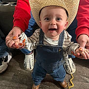 Kamdyn is registered to the contest to win money with this photo: toddler, child, cowboy_hat, denim_overalls, smiling, indoor, wooden_floor, toy_snake, colorful_sock, hands, nail_art, red_sweatshirt, adult, shoes, striped_shirt, happy, person, footwear, supporting, playful