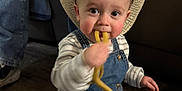 Kamdyn joined the competition — help win amazing prizes! toddler, child, cowboy_hat, denim_overalls, snack, floor, indoor, person, sitting, curly_food, cute, baby, clothing, jeans, striped_shirt, face, chewing, hat, footwear, background