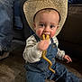 toddler, child, cowboy_hat, denim_overalls, snack, floor, indoor, person, sitting, curly_food, cute, baby, clothing, jeans, striped_shirt, face, chewing, hat, footwear, background