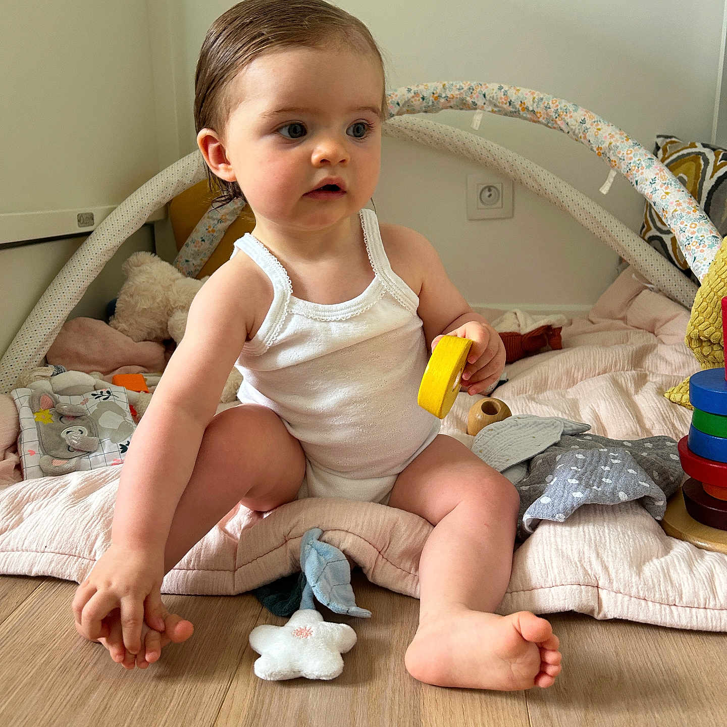 Lou a rejoint le concours — aidez-le/la à gagner de superbes lots ! baby, child, curious, cushion, face, foot, hair, hand, home, indoor, natural_light, pillow, playmat, plush_toy, sitting, soft_toys, toy, white_tank_top, wooden_floor, yellow_toy