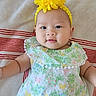 baby, infant, child, face, headband, flower, yellow, dress, floral, pattern, blanket, striped, smile, cute, lying_down, skin, arm, portrait, indoors, person