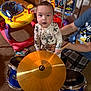baby, child, toy_drum, cymbal, pajamas, adult, supporting_hand, carpet, indoor, toys, walker, box, shelf, plaid_pants, blue_shirt, music, playtime, curious, expression, floor
