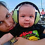 baby, adult, headphones, outdoor, stadium, bleachers, tshirt, taco, child, person, holding, face, blue_sky, casual_clothing, seated_people, portrait, daytime, expressionless, closeup, event