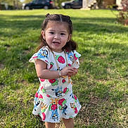 Leilani is registered to the contest to win money with this photo: toddler, child, smiling, dress, cartoon_print, grass, outdoor, sunlight, accessories, bracelet, sandals, gold_chain, happy, standing, nature, person, young_child, greenery, playful, summer