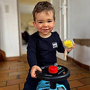 Owen a rejoint le concours — aidez-le/la à gagner de superbes lots ! toddler, child, toy_car, indoor, smiling, black_clothing, steering_wheel, yellow_toy, short_hair, floor_tiles, playful, happy, person, young_child, casual_clothing, childhood, cute, home_interior, portrait, fun