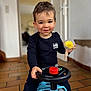 toddler, child, toy_car, indoor, smiling, black_clothing, steering_wheel, yellow_toy, short_hair, floor_tiles, playful, happy, person, young_child, casual_clothing, childhood, cute, home_interior, portrait, fun