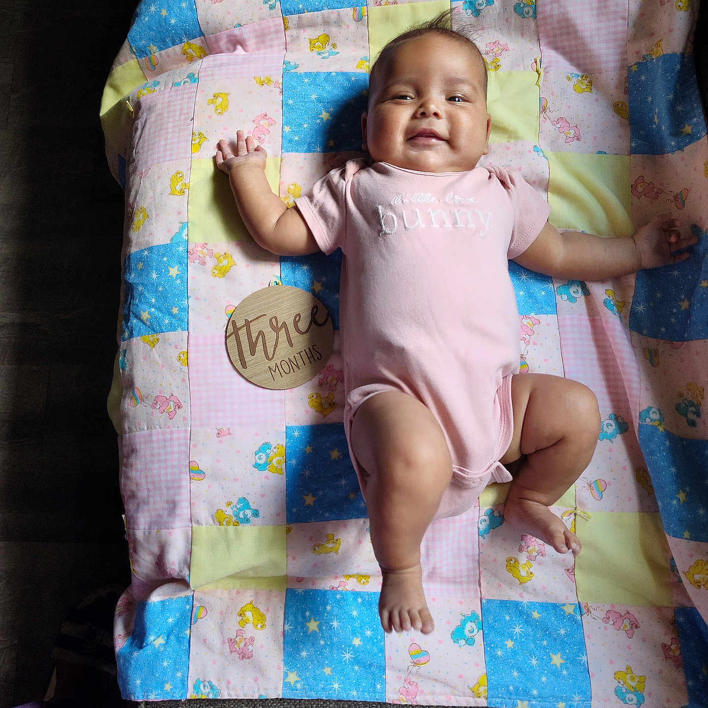 Glorianna joined the competition — help win amazing prizes! baby, blanket, care_bears, child, cute, fabric, floor, happy, indoors, infant, lying_down, patchwork_blanket, person, pink_onesie, skin, small_child, smiling, soft_light, three_months, wooden_sign
