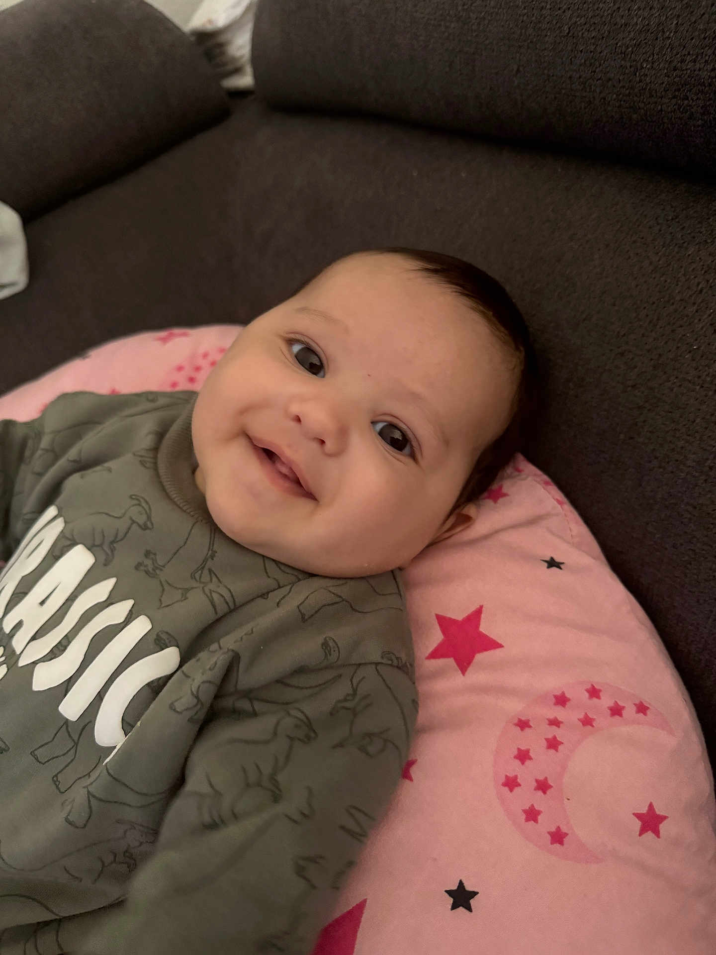 Naseem a rejoint le concours — aidez-le/la à gagner de superbes lots ! baby, smiling, face, couch, pink_cushion, stars, moon, green_sweatshirt, dinosaur_pattern, infant, child, apparel, indoor, happy, portrait, closeup, cozy, young_child, person, relaxing